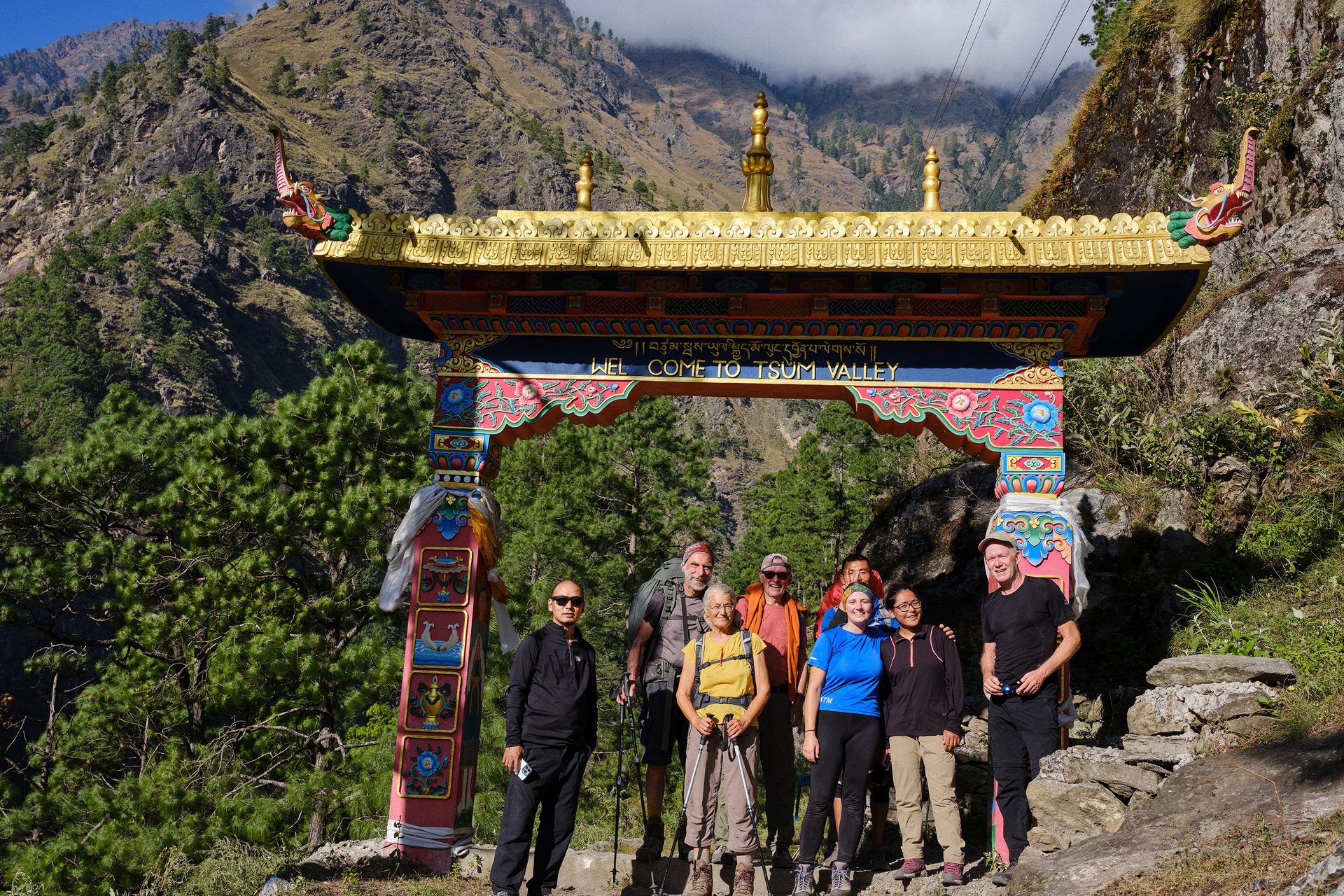 Trekkers beneath the ornate golden gateway that marks the entrance to Tsum Valley, forested ridges behind